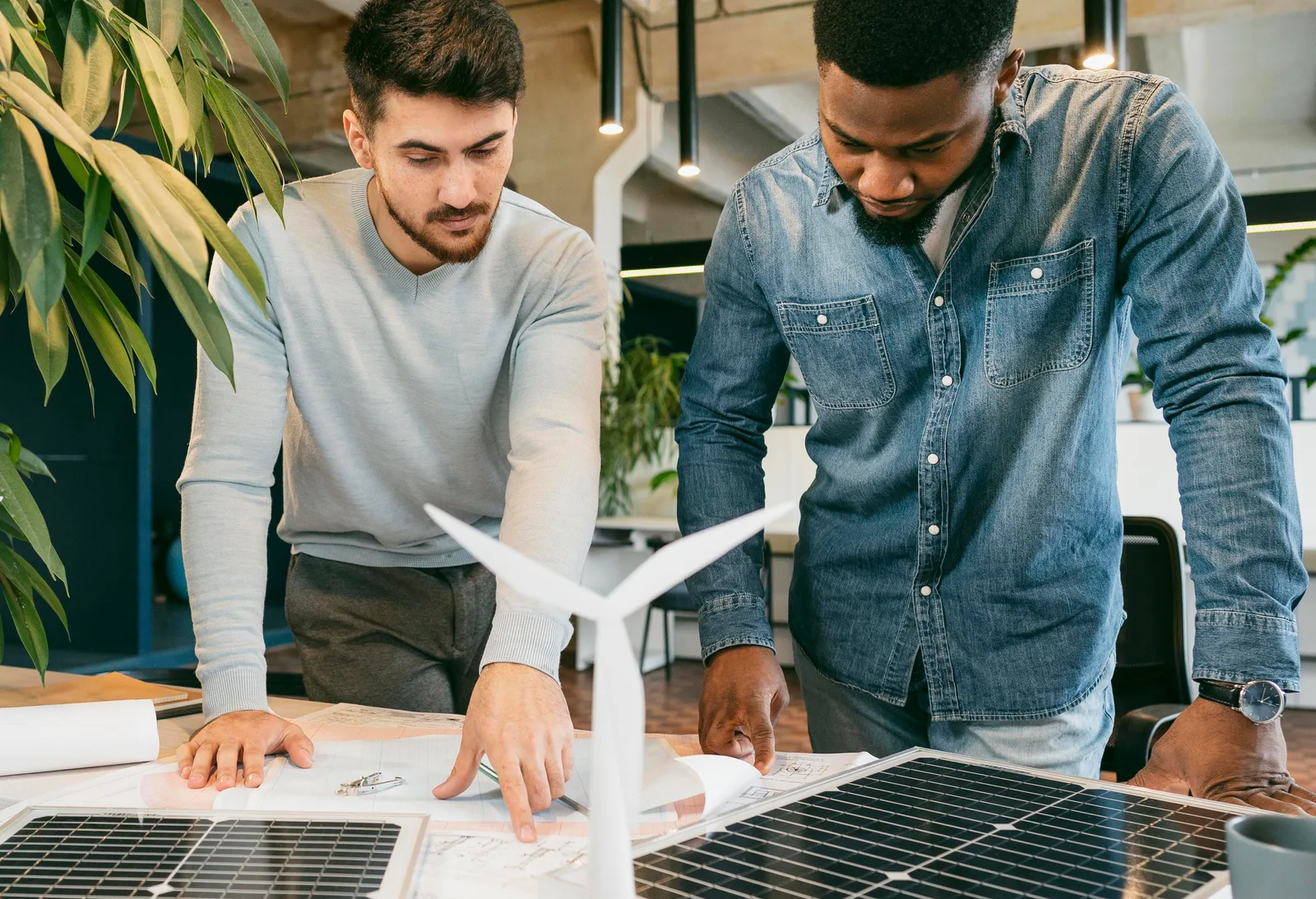 Dois profissionais analisam projetos técnicos sobre uma mesa com painéis solares e maquete de turbina eólica