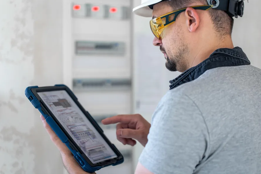 Eletricista de capacete e óculos de proteção analisa dados em um tablet diante do quadro elétrico