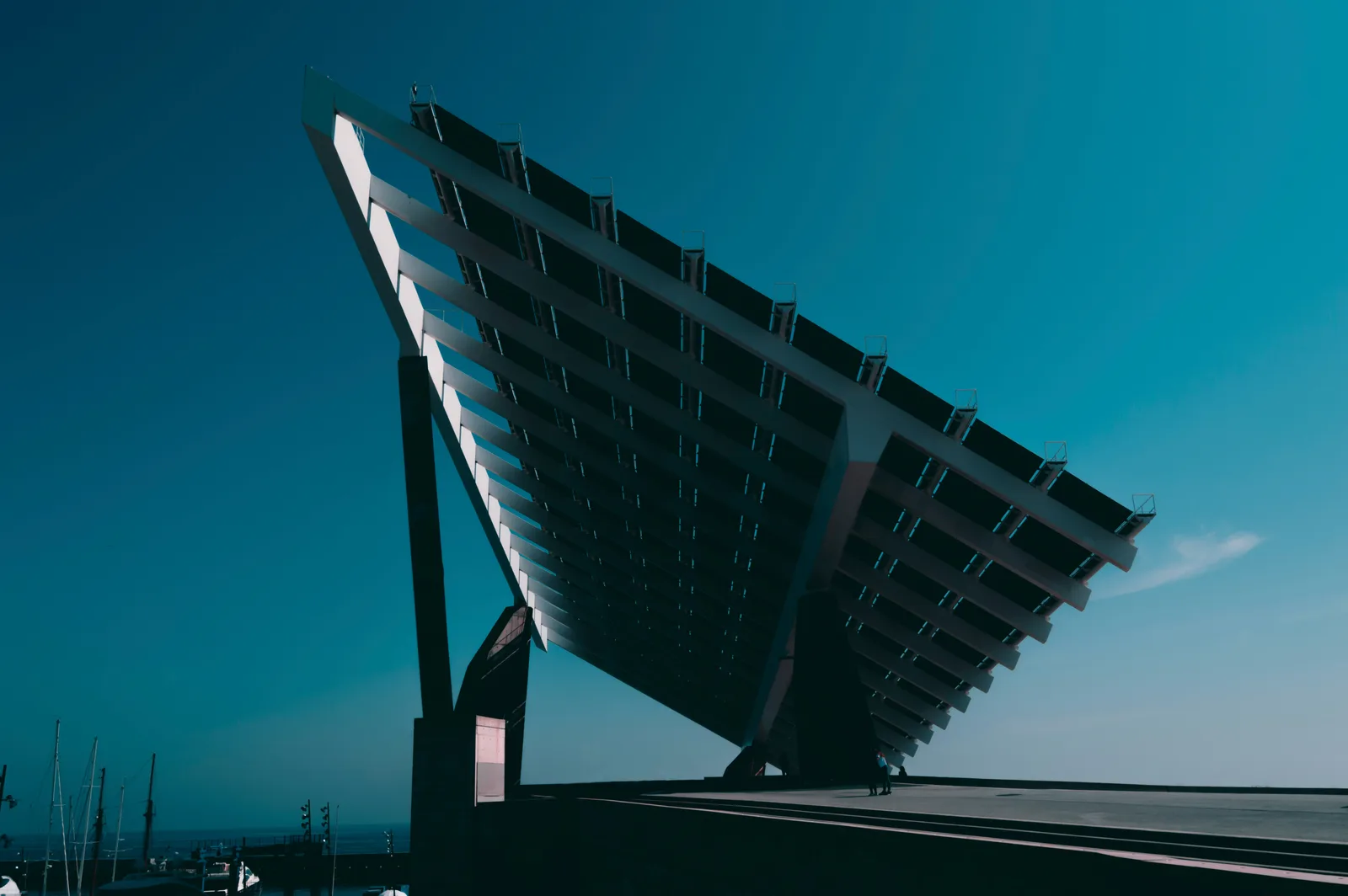 Grande estrutura inclinada de painéis solares contra o céu azul, com mastros de uma marina ao fundo