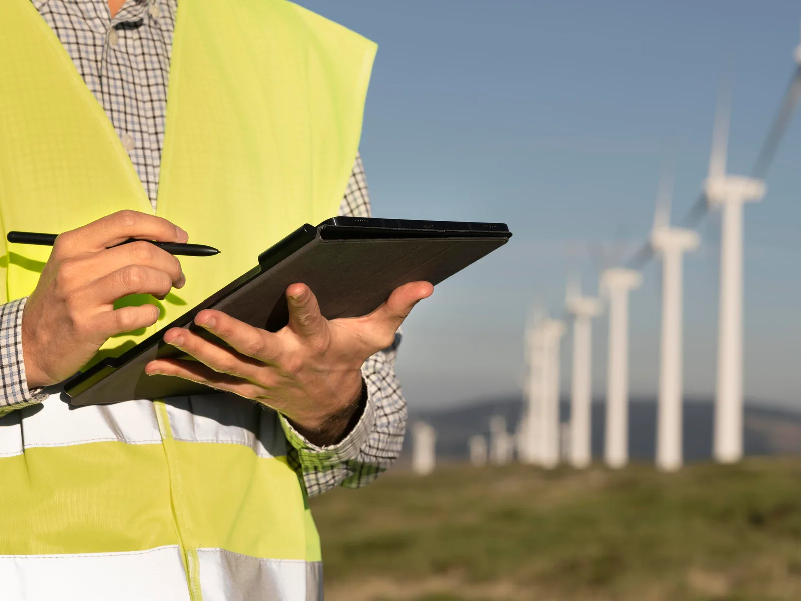 Técnico com colete de alta visibilidade faz anotações num tablet diante de uma fileira de aerogeradores