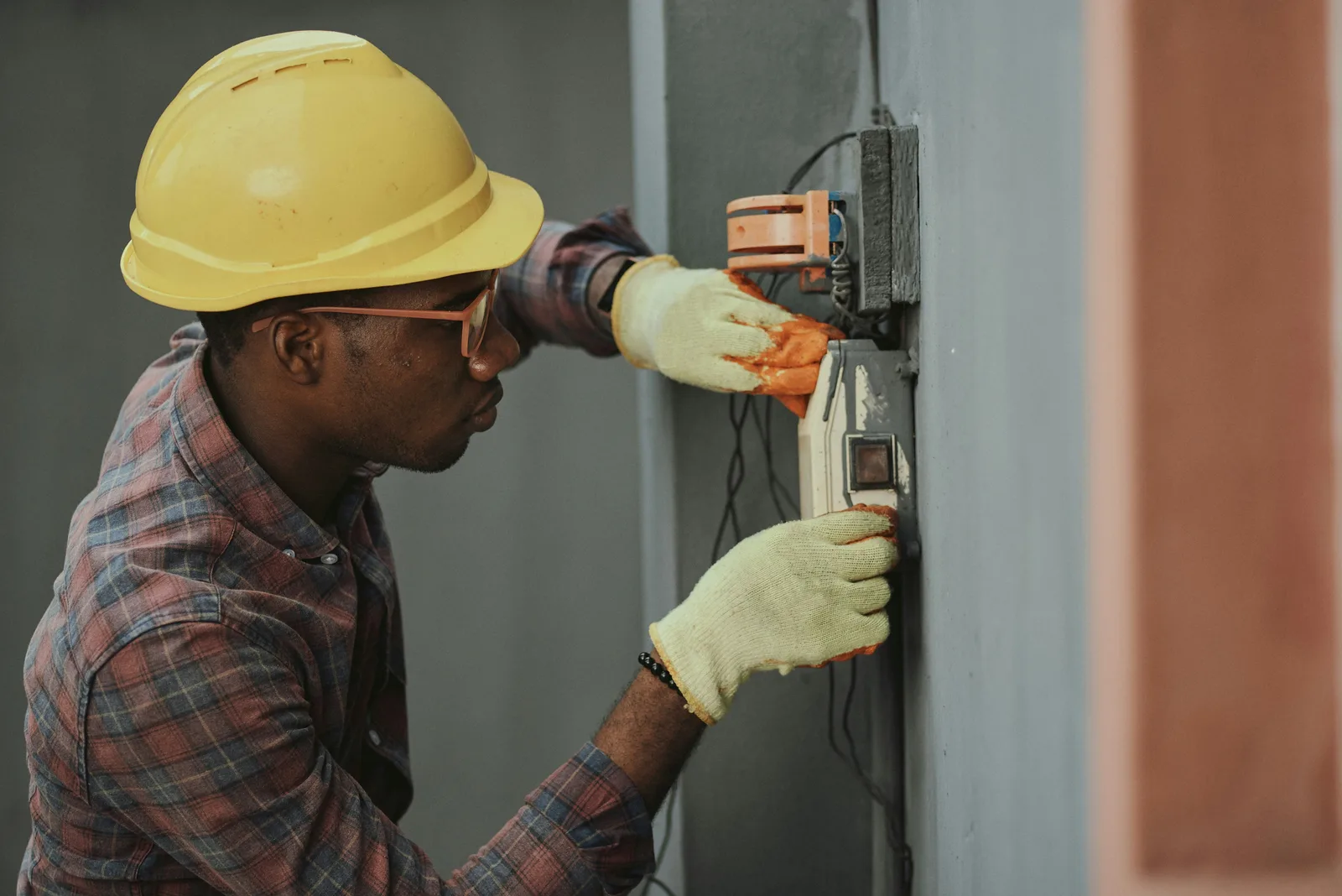Técnico com capacete amarelo e luvas verifica um dispositivo elétrico instalado na parede