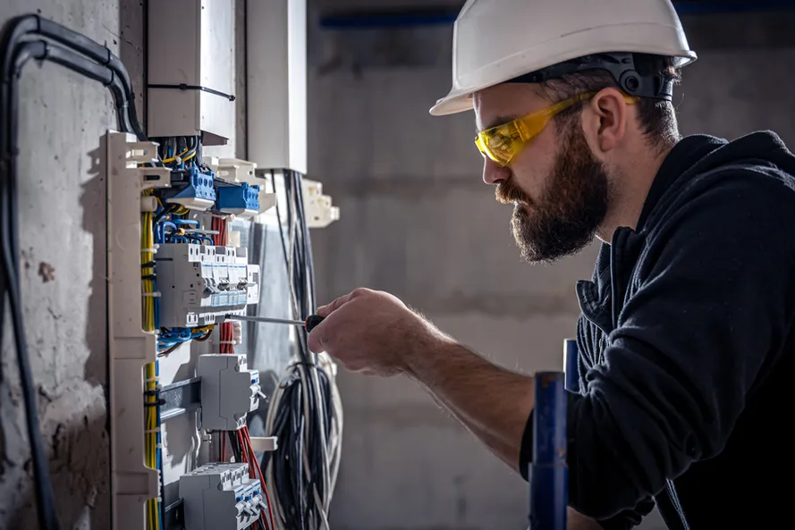 Eletricista de capacete e óculos de proteção aperta conexões em um quadro elétrico com chave de fenda