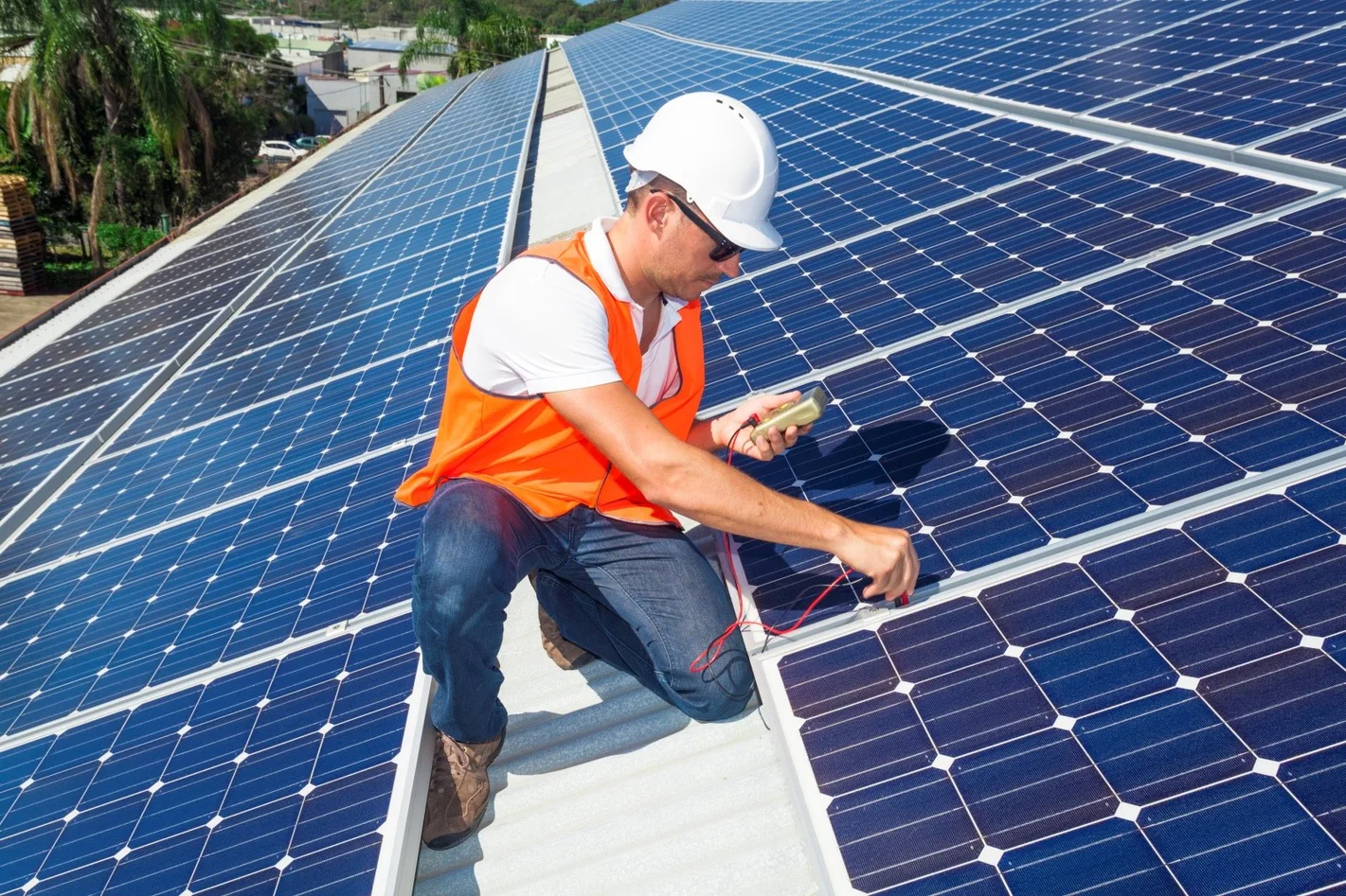 Técnico de capacete e colete laranja mede painéis solares com multímetro em um telhado ensolarado