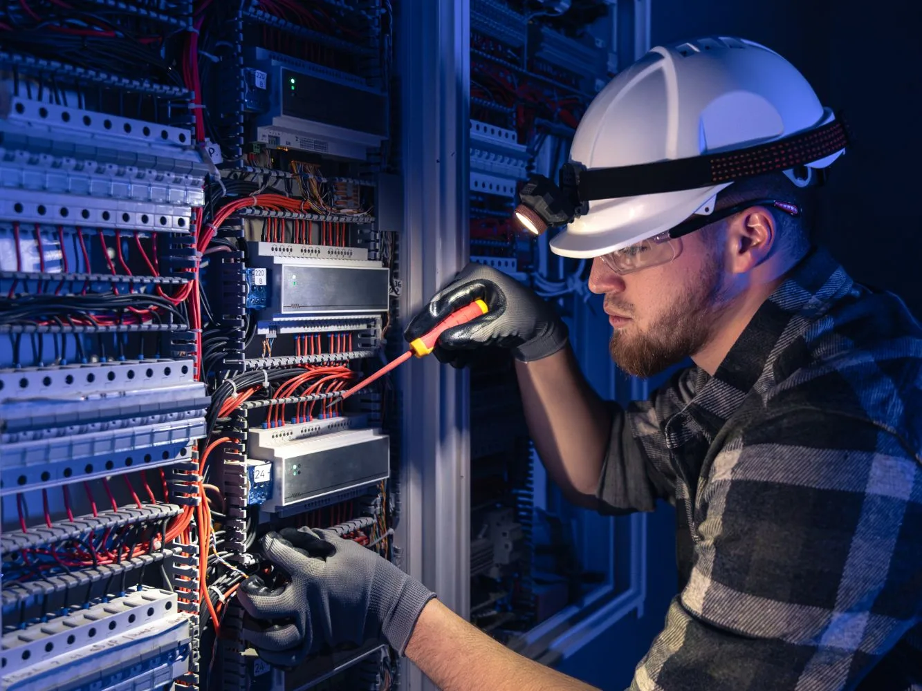 Eletricista de capacete e lanterna na cabeça fazendo manutenção em um quadro elétrico com chave de fenda