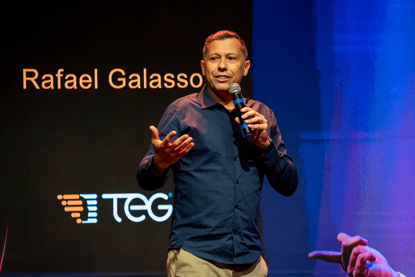 Rafael Galasso ao microfone num evento da TEG, com o seu nome e o logótipo TEG no ecrã ao fundo